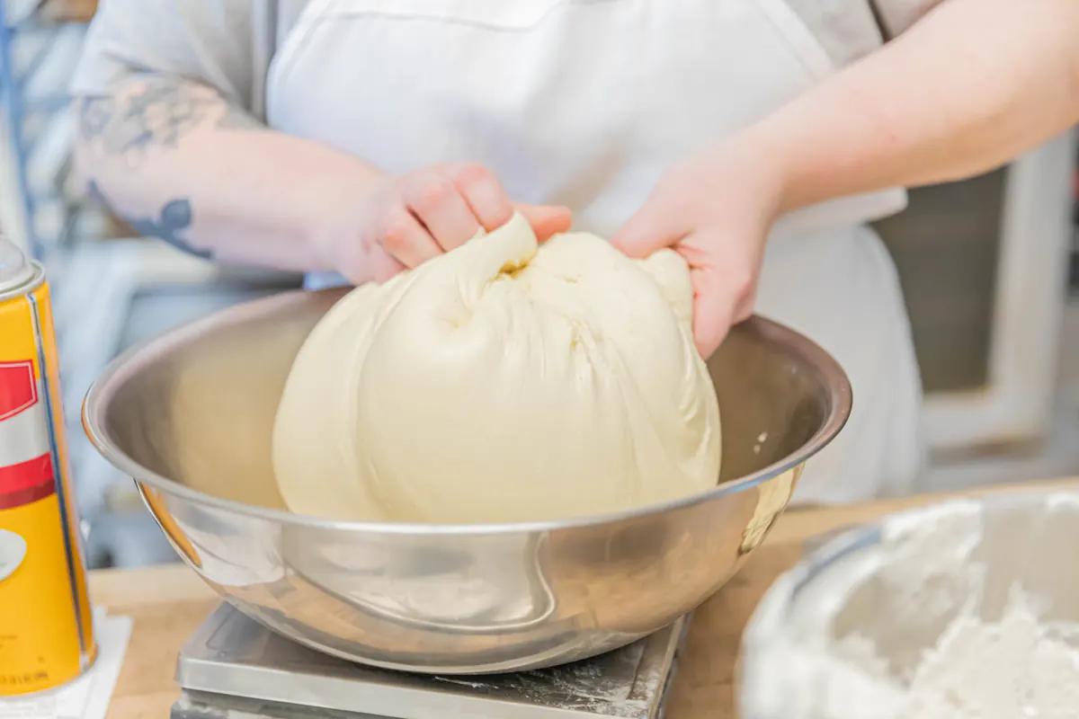 A baker lifting a smooth ball of dough from a steel bowl